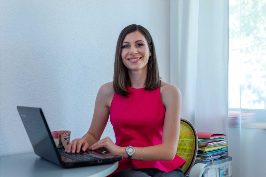 Karolina sjedi za laptopom u crvenoj majici i gleda u kameru